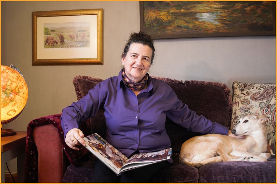 Marina at home in Scotland, Oman travel book on her lap, a globe showing the Arabian Peninsula beside her, and her dog Ramlah resting on the sofa